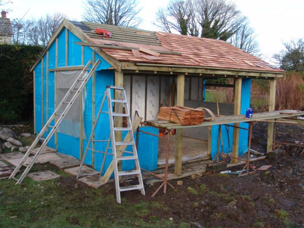 Timber frame of a garden room wrapped in blue membrane, with the shingle roof going on
